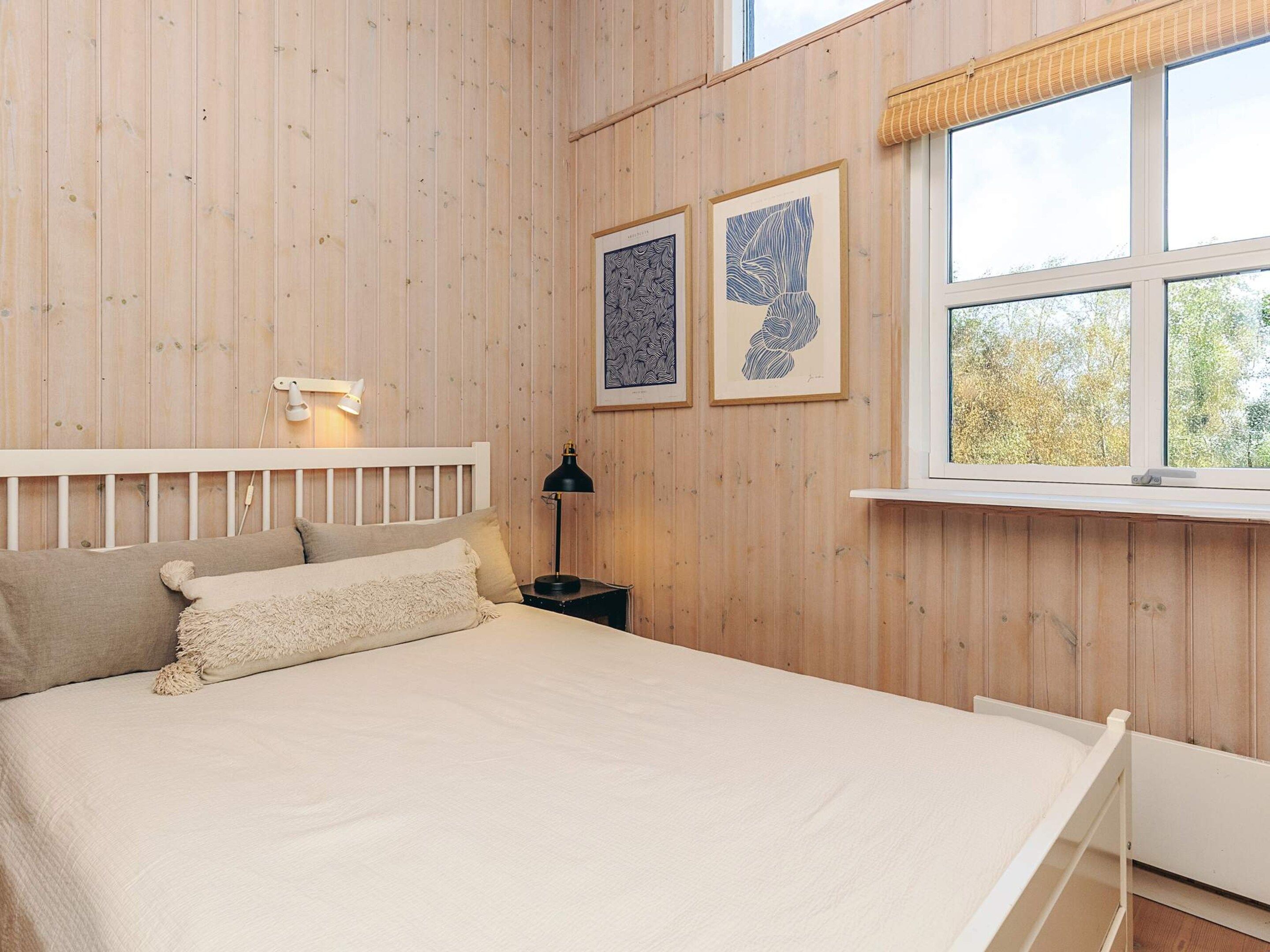 A bedroom with a wooden paneled wall, a bed with a white headboard, a bedside lamp, and two framed artworks.
