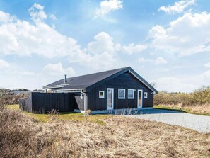 A single-story, dark-colored wooden house with a sloped roof, a front porch, and a driveway leading to the entrance. - 4 Star Holiday Home in Hirtshals (Hirtshals)