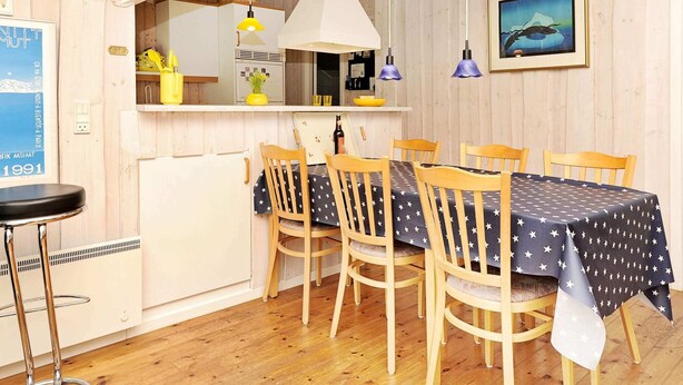 Comedor con una mesa lista para cuatro personas, sillas de madera, un taburete de bar, área de cocina con refrigerador blanco y un cuadro en la pared.