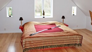 A bedroom with a sloped ceiling, a bed with a patterned bedspread, two bedside lamps, and a window with a view of greenery.