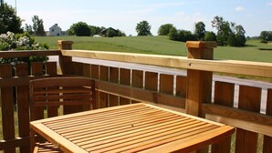A wooden deck with a table and chairs, overlooking a grassy field and a white building.