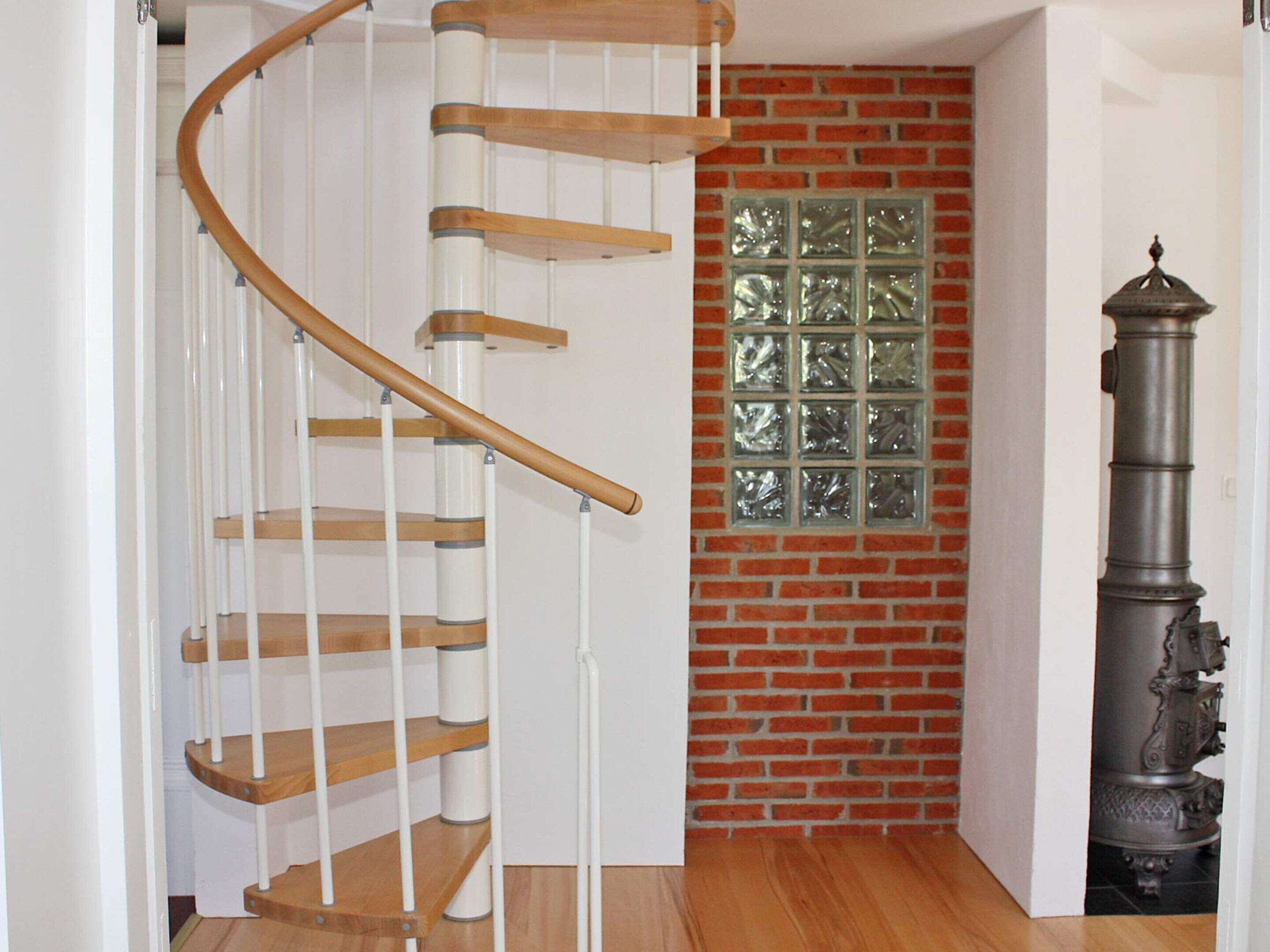 A spiral staircase with wooden steps and white railings, a brick wall with a decorative window, and a tall, ornate metal object.
