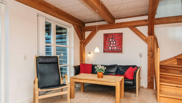 A room with a wooden ceiling, a black leather chair, a wooden table, a black sofa with red cushions, and a wooden staircase.