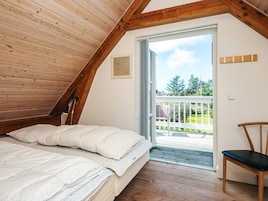 A bedroom with a sloped ceiling, a bed, a chair, and a balcony with a view of greenery.