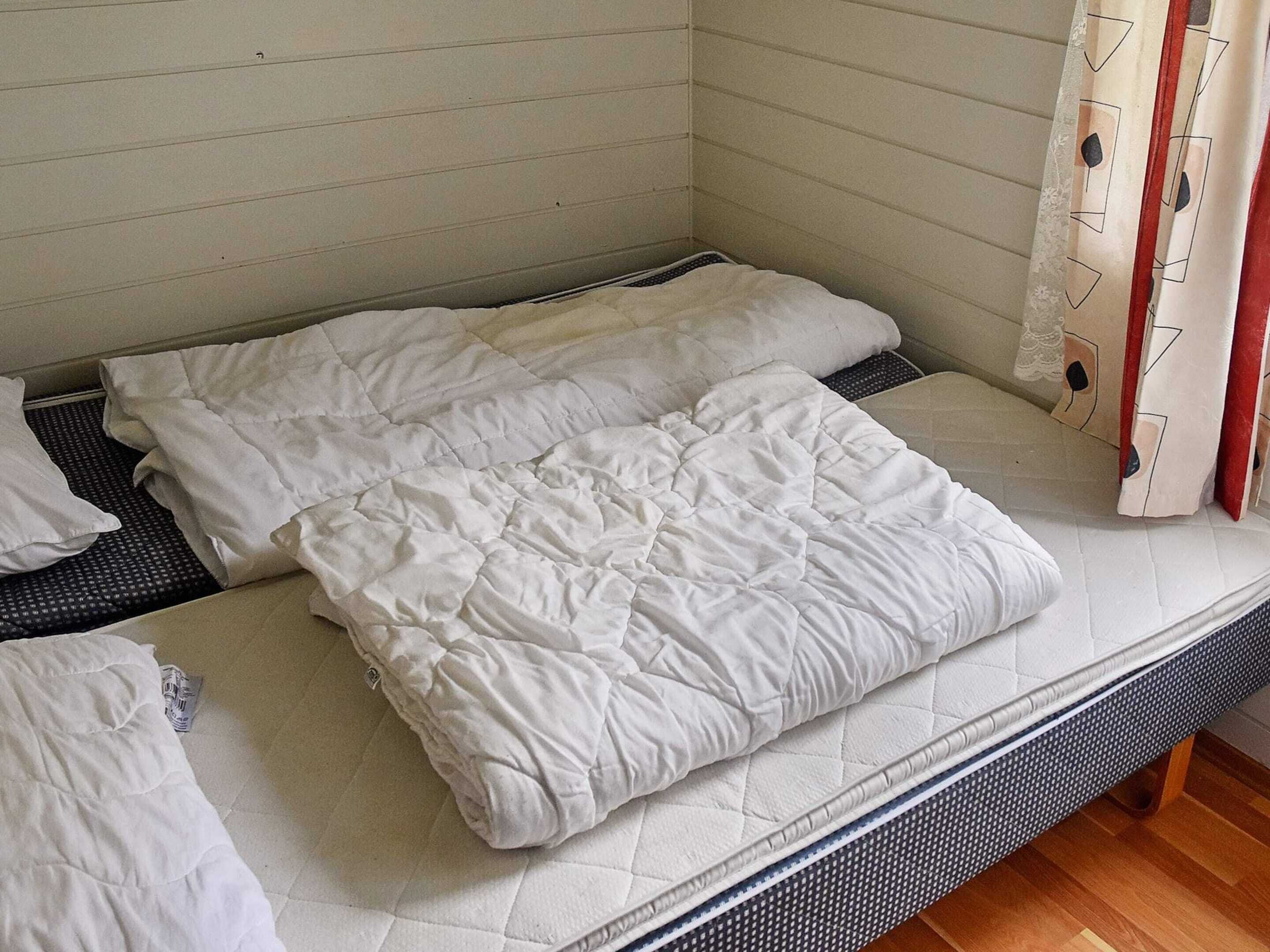 A single bed with a white mattress and bedding, a wooden floor, and a white wall with a window.