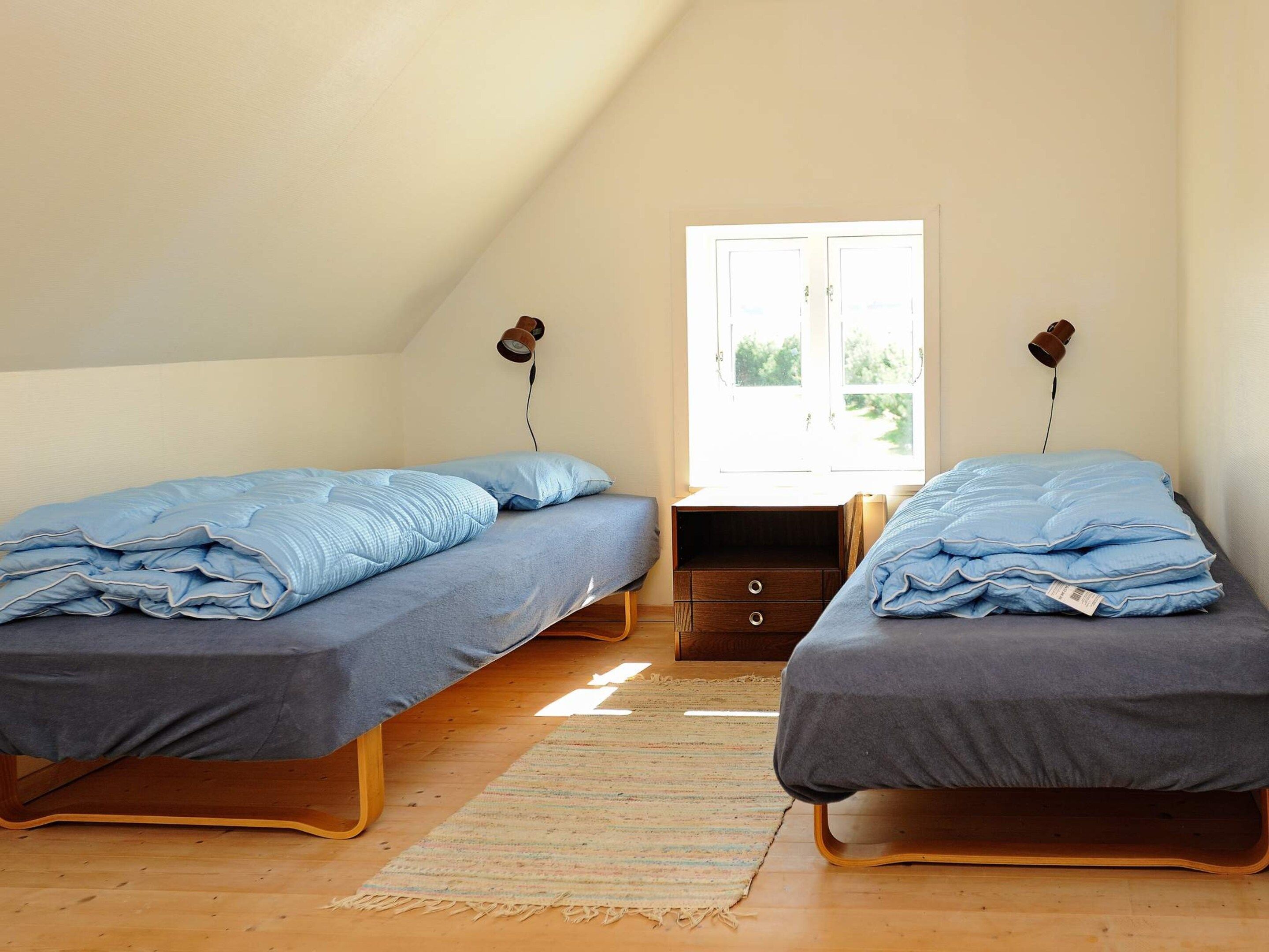 Quarto com duas camas, uma pequena mesa de cabeceira de madeira e uma janela com luz natural.