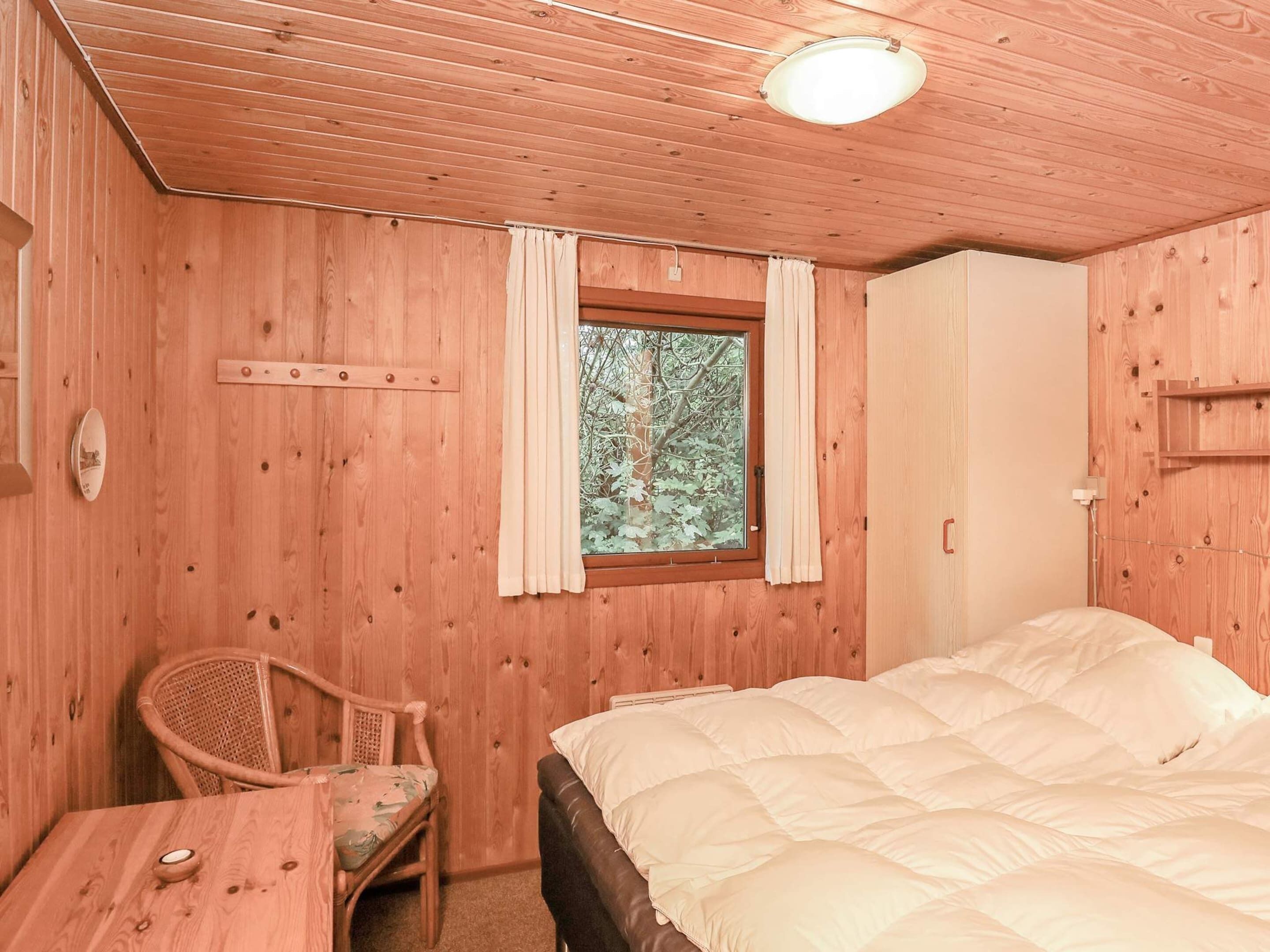 A wooden cabin room with a bed, a chair, a table, and a window with curtains.