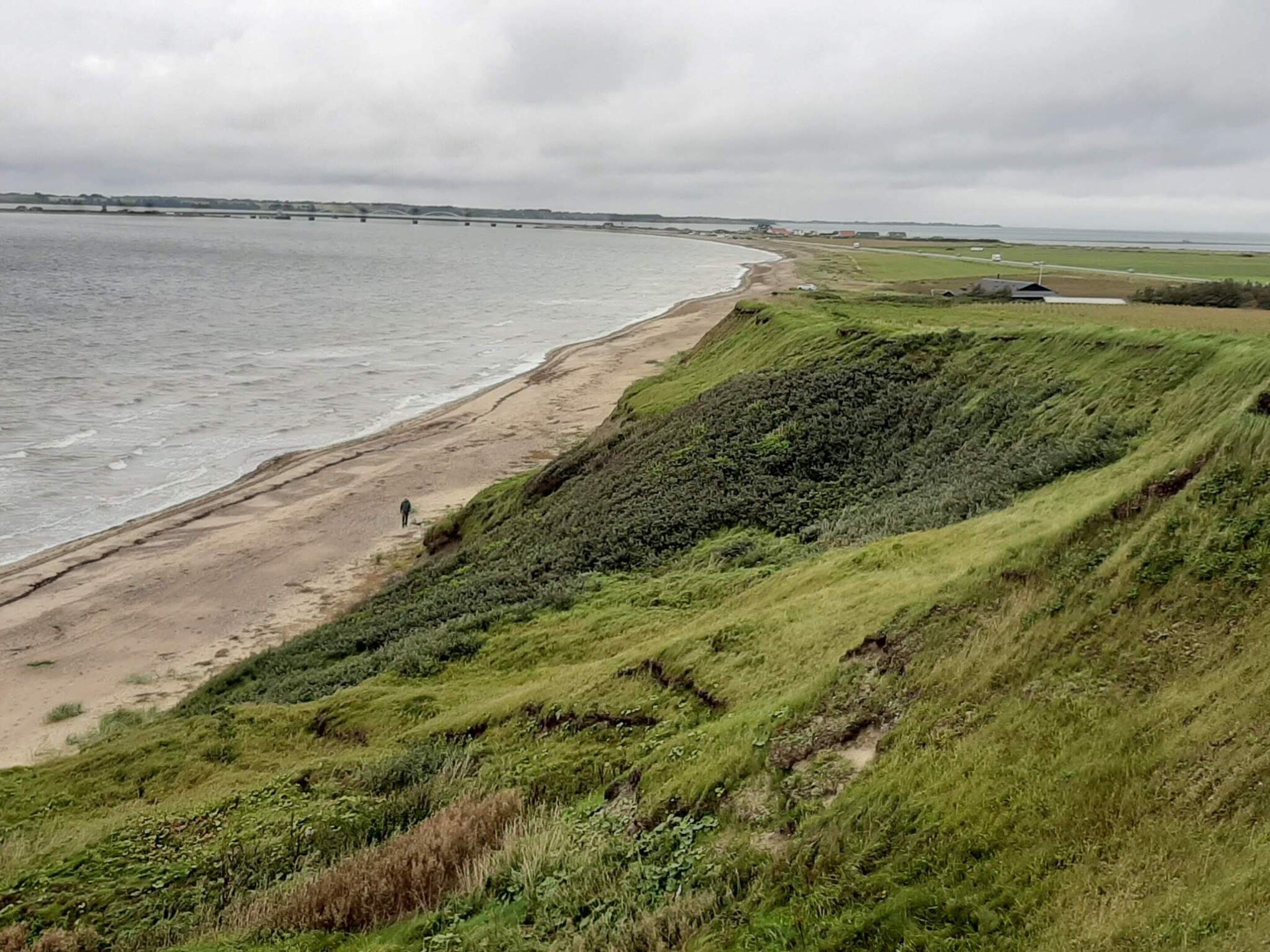 A coastal landscape with a beach, grassy terrain, and a distant bridge.