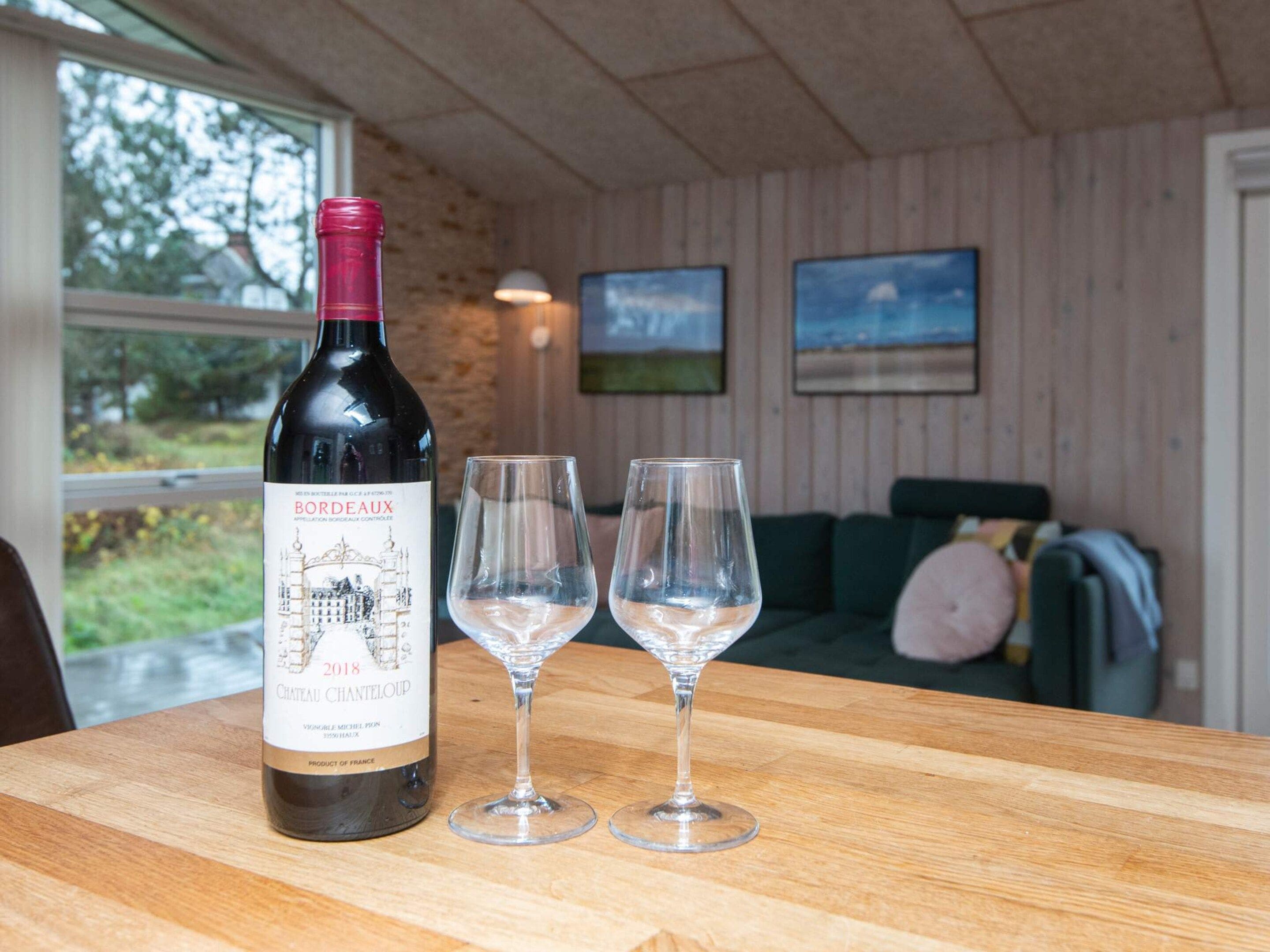 A bottle of wine and two glasses on a wooden table in a room with a view of greenery outside.