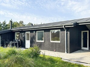 Una cabaña moderna de madera oscura con techo plano, ventanas amplias y un patio con mobiliario exterior.