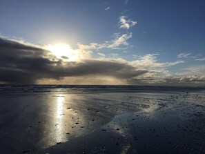 A beach with wet sand reflecting sunlight, a cloudy sky, and the ocean in the background.