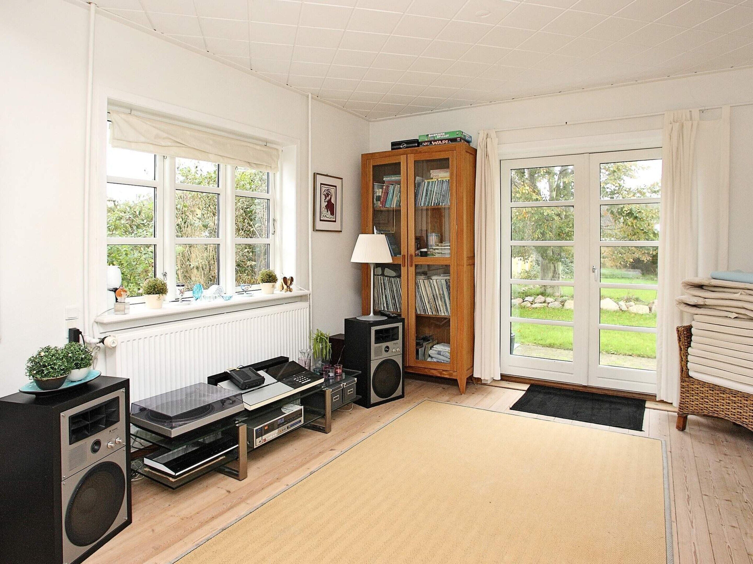 Une pièce avec un radiateur, un haut-parleur noir, un meuble en bois rempli de livres et une fenêtre donnant sur un paysage verdoyant.