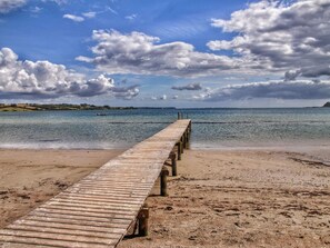 Un moll de fusta que s'endinsa en aigües tranquil·les, amb una platja sorrenca i les muntanyes a la distància.