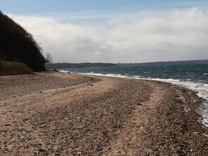 A pebbly beach with waves, a distant shoreline, and overcast sky.