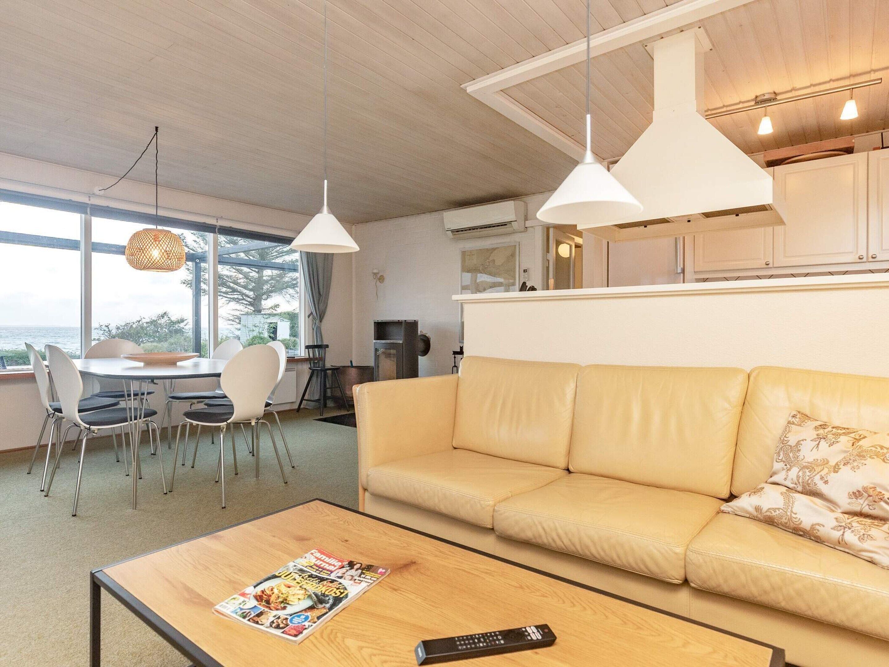 A living room with a sofa, dining table, chairs, a coffee table with a magazine and remote control, pendant lights, and a view through large windows.