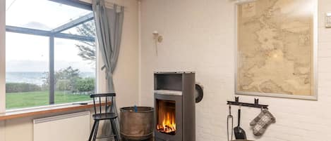A room with a wood-burning stove, a map on the wall, a window with a view of the sea, and a chair.