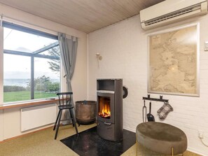 A room with a wood-burning stove, a map on the wall, a window with a view of the sea, and a chair.