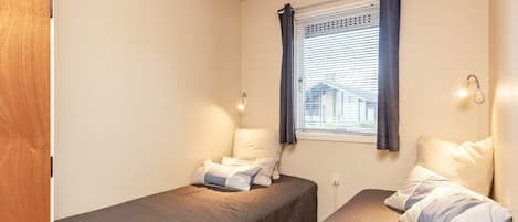 Une petite chambre avec deux lits simples, une fenêtre avec des rideaux et une lampe fixée au mur.