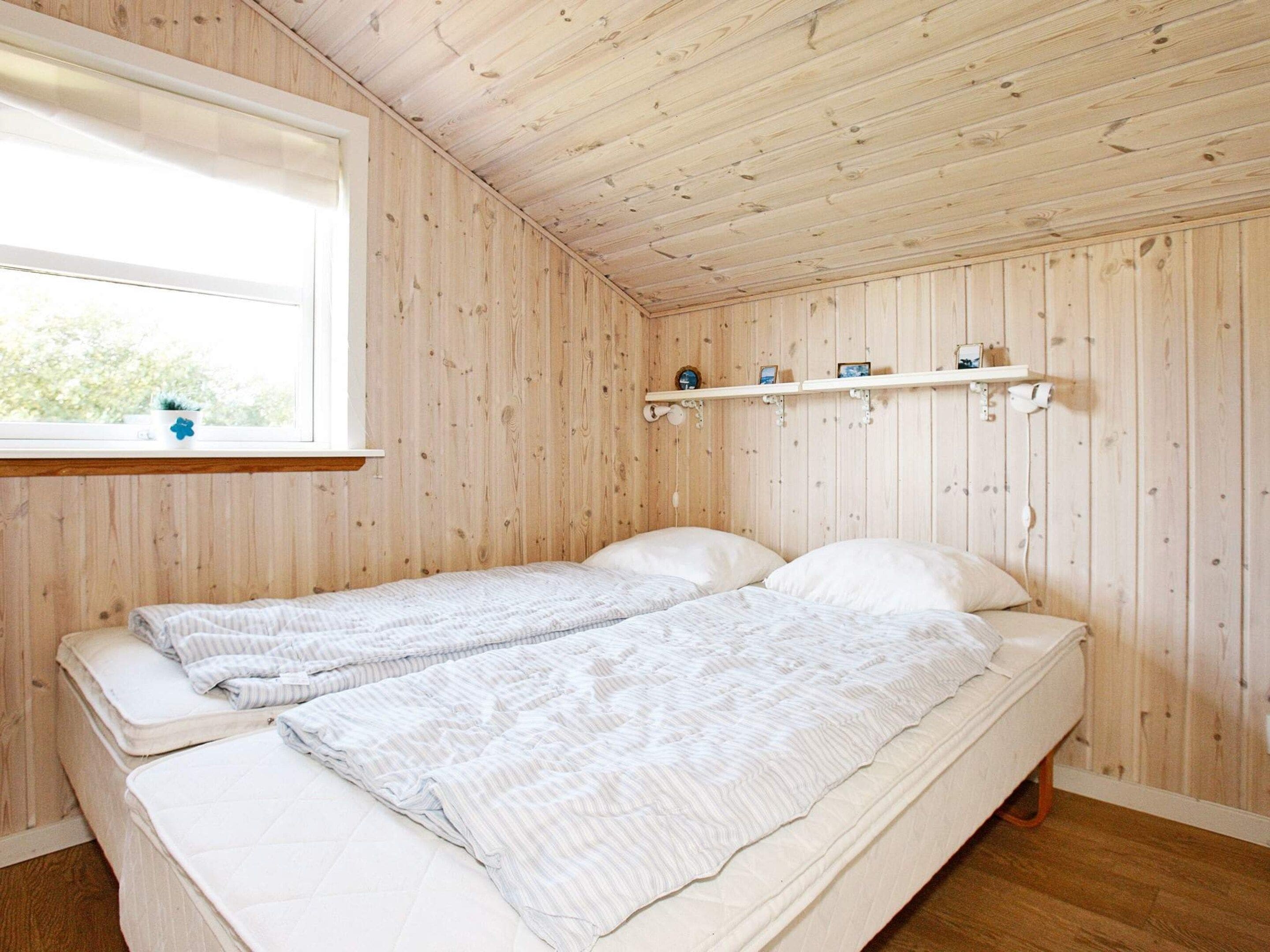 A room with wooden walls and a single bed with two pillows. There is a window with a view of greenery outside.