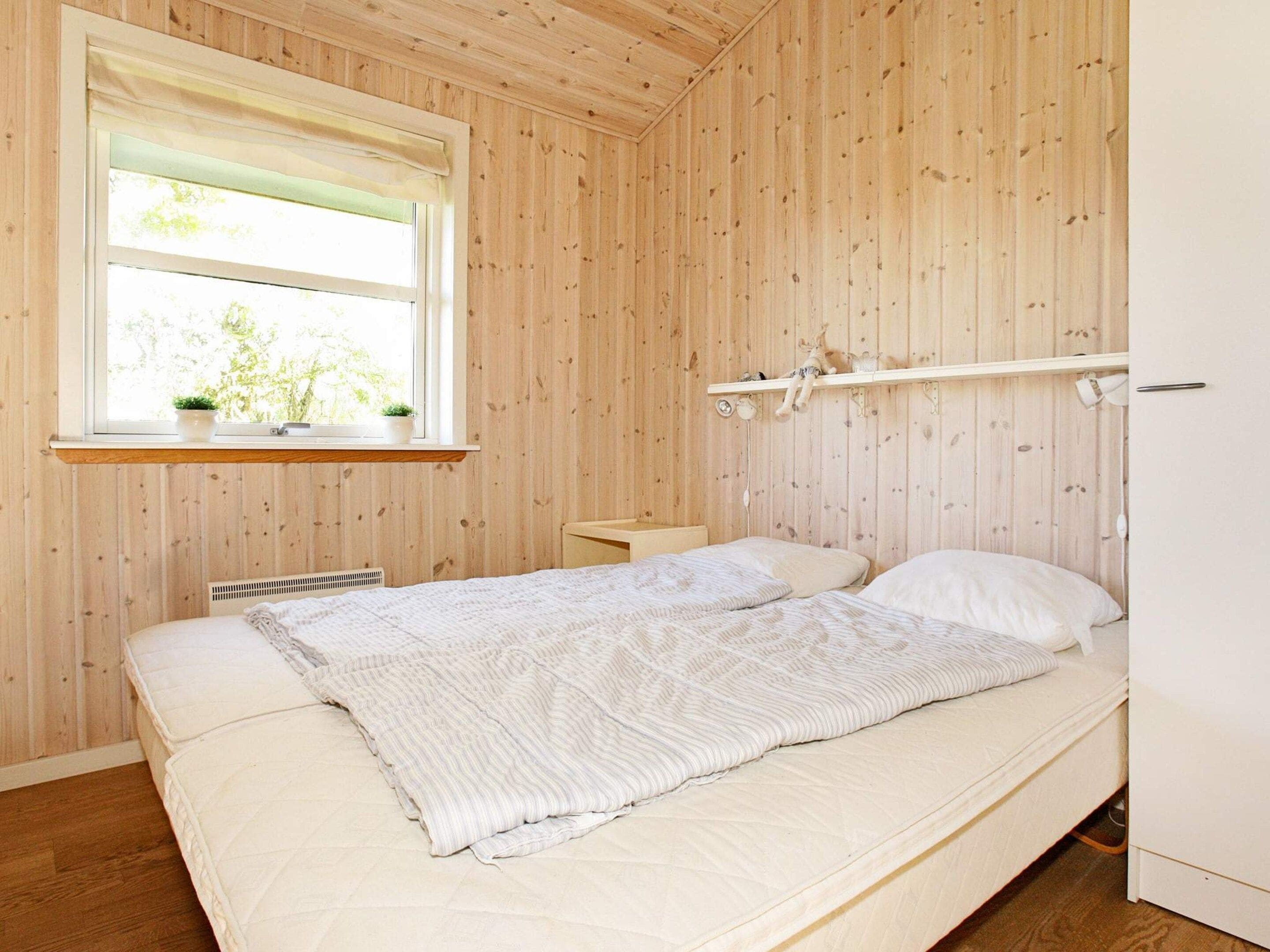 A room with a bed, a window with a view, a shelf with a plant, and a wall-mounted lamp.