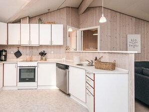 A modern kitchen with white cabinets, a dishwasher, and a built-in oven. There is a wooden wall paneling, a hanging light fixture, and a basket on the countertop.