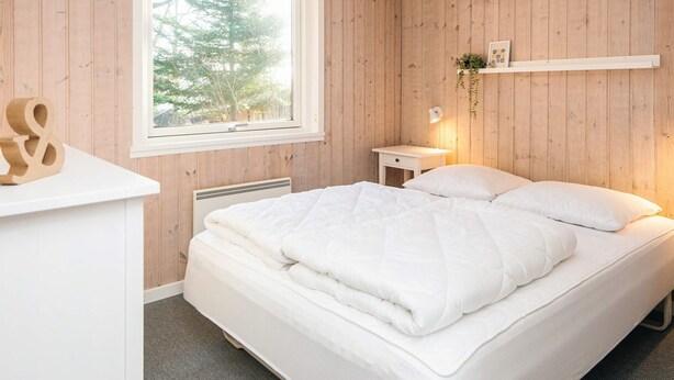 A bedroom with a wooden paneled wall, a bed with white bedding, a bedside table with a lamp, and a window with a view of trees. - 8 Person Holiday Home in Borkop (Borkop)