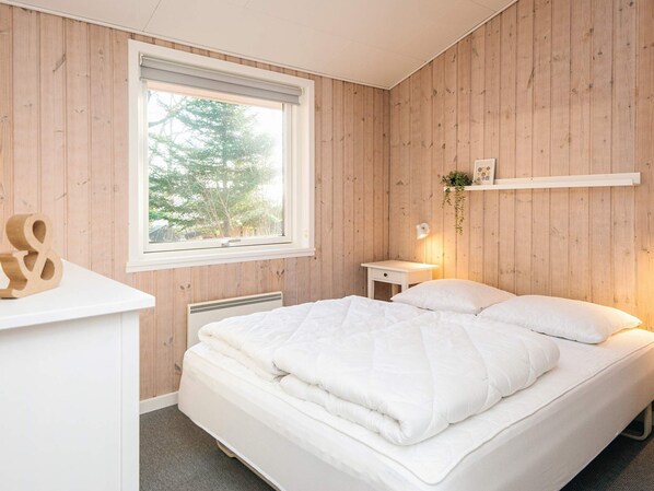 A bedroom with a wooden paneled wall, a bed with white bedding, a bedside table with a lamp, and a window with a view of trees.