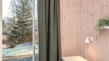 A room with a wooden wall, a small white table, a lamp, and a window with a curtain.