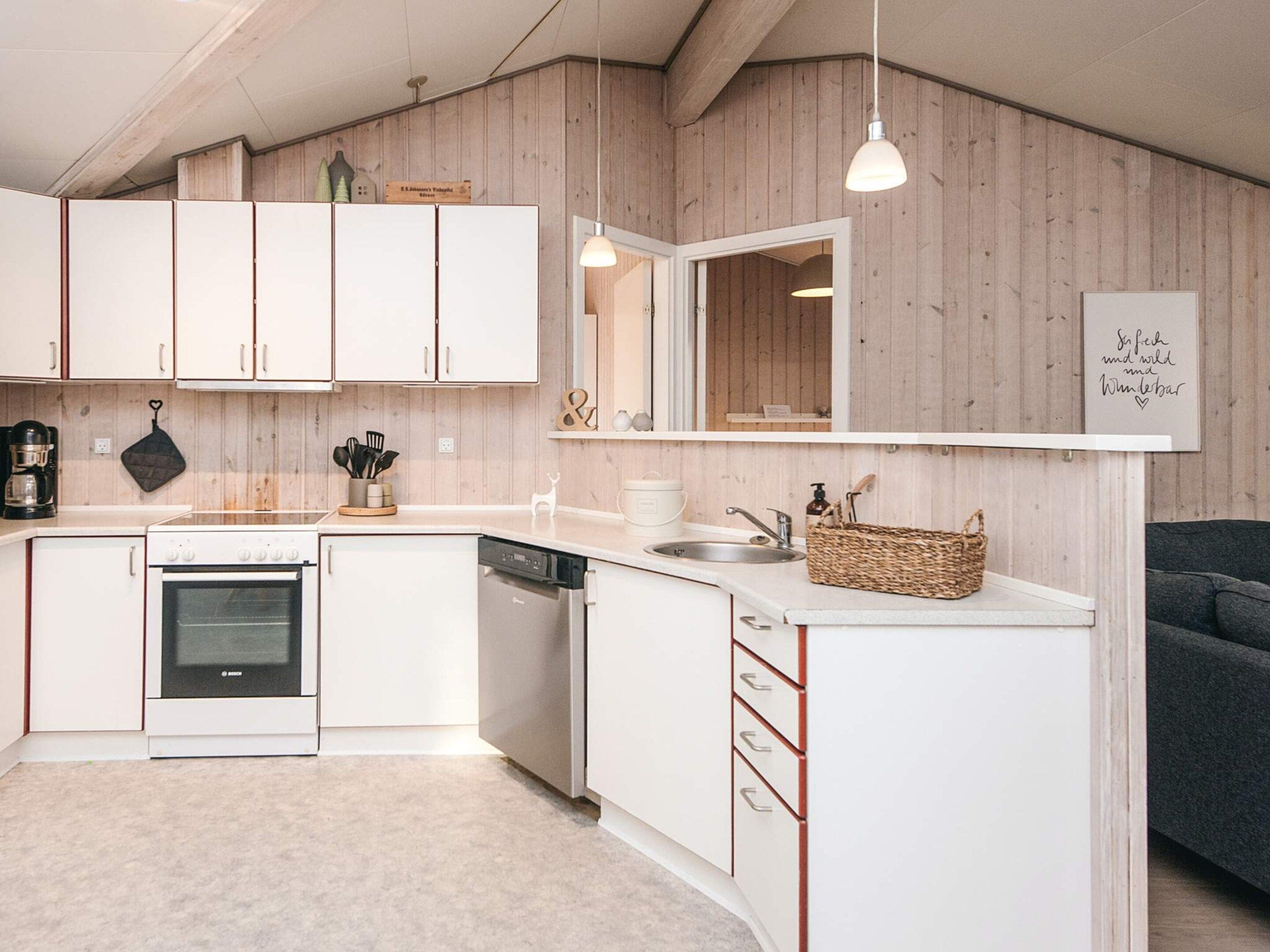 Cozinha moderna com armários brancos, lava-louças e forno embutido. Há uma parede de madeira com uma prateleira acima da pia e um lustre.