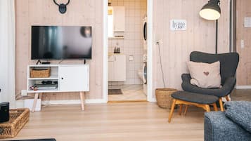 Un salón con televisor de pantalla plana, un sillón gris oscuro con un cojín con forma de corazón, un mueble blanco y un cartel que dice 'hygg'.