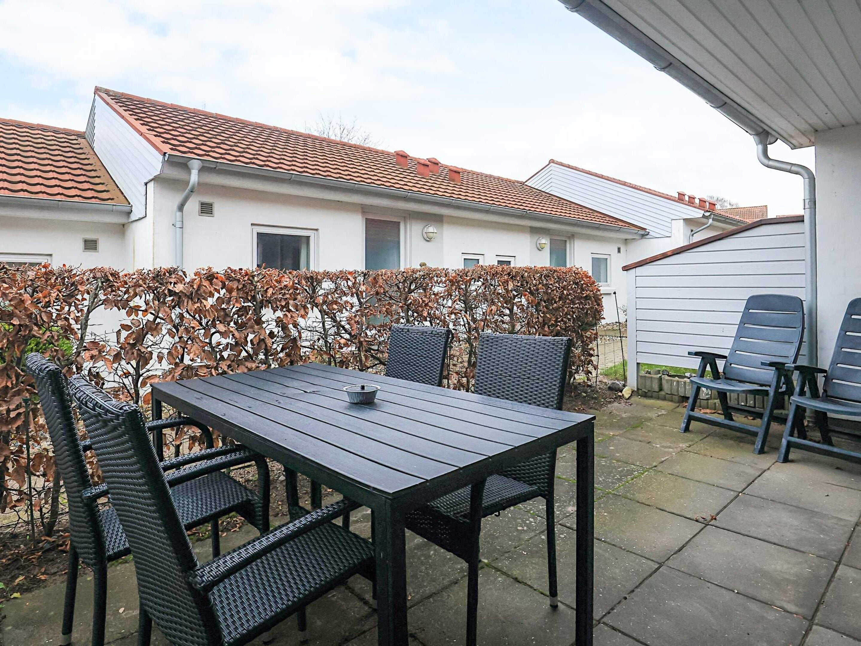 Une terrasse extérieure avec une table et des chaises, une haie et une maison en arrière-plan.