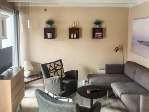 Un salon moderne avec un canapé, des fauteuils, une table basse et un téléviseur à écran plat. Des étagères murales accueillent des objets décoratifs et un tableau orne le mur.