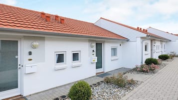 A row of white buildings with red-tiled roofs, small windows, and a paved walkway with gravel and plants.