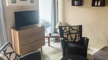 A living room with a flat-screen TV on a wooden cabinet, two black armchairs, a small round table with a remote control, and a glass coffee table.