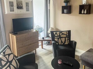 A living room with a flat-screen TV on a wooden cabinet, two black armchairs, a small round table with a remote control, and a glass coffee table.