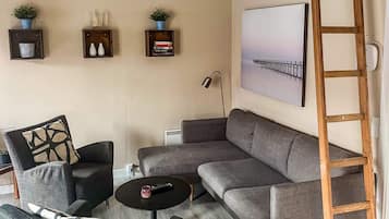 Un salon avec un canapé gris, deux fauteuils, une table basse et une échelle appuyée contre le mur. On y trouve trois étagères ornées d’objets décoratifs et un tableau au-dessus du canapé.