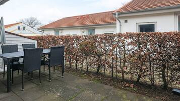 An outdoor patio area with a table and chairs, a hedge, and a building with a tiled roof in the background.