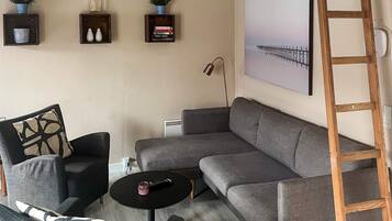 Un salon moderne avec un canapé d’angle gris, une petite table ronde et deux fauteuils. Trois étagères murales sont ornées d’objets décoratifs et d’un tableau encadré. Une échelle en bois est appuyée contre le mur.
