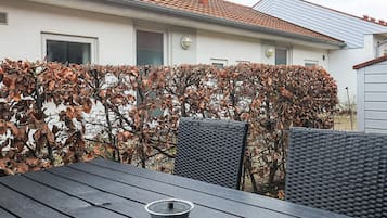 A patio with a table and chairs, a small bowl, and a house with a tiled roof and a hedge.