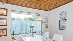 A dining area with a table covered in a patterned tablecloth, white chairs, a hanging light fixture, and a clock on the wall.