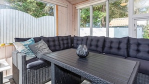 A covered outdoor seating area with a dark-colored sofa and a table, surrounded by wooden walls and large windows.