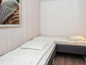 Uma cama pequena e bem arrumada com roupa de cama branca em um quarto com paredes de madeira clara e teto branco sem adornos.