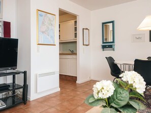 En stue med fjernsyn, et spiseområde med bord og stole, et køkken med skabe og vask, samt en potteplante med hvide blomster.