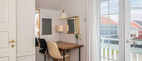A room with a wooden desk, a chair, a lamp, a mirror, and a view of the outside through a glass door.