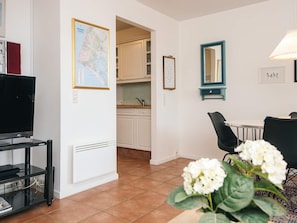 A modern living room with a television, a dining area with a table and chairs, a kitchen in the background, and a potted plant with white flowers.