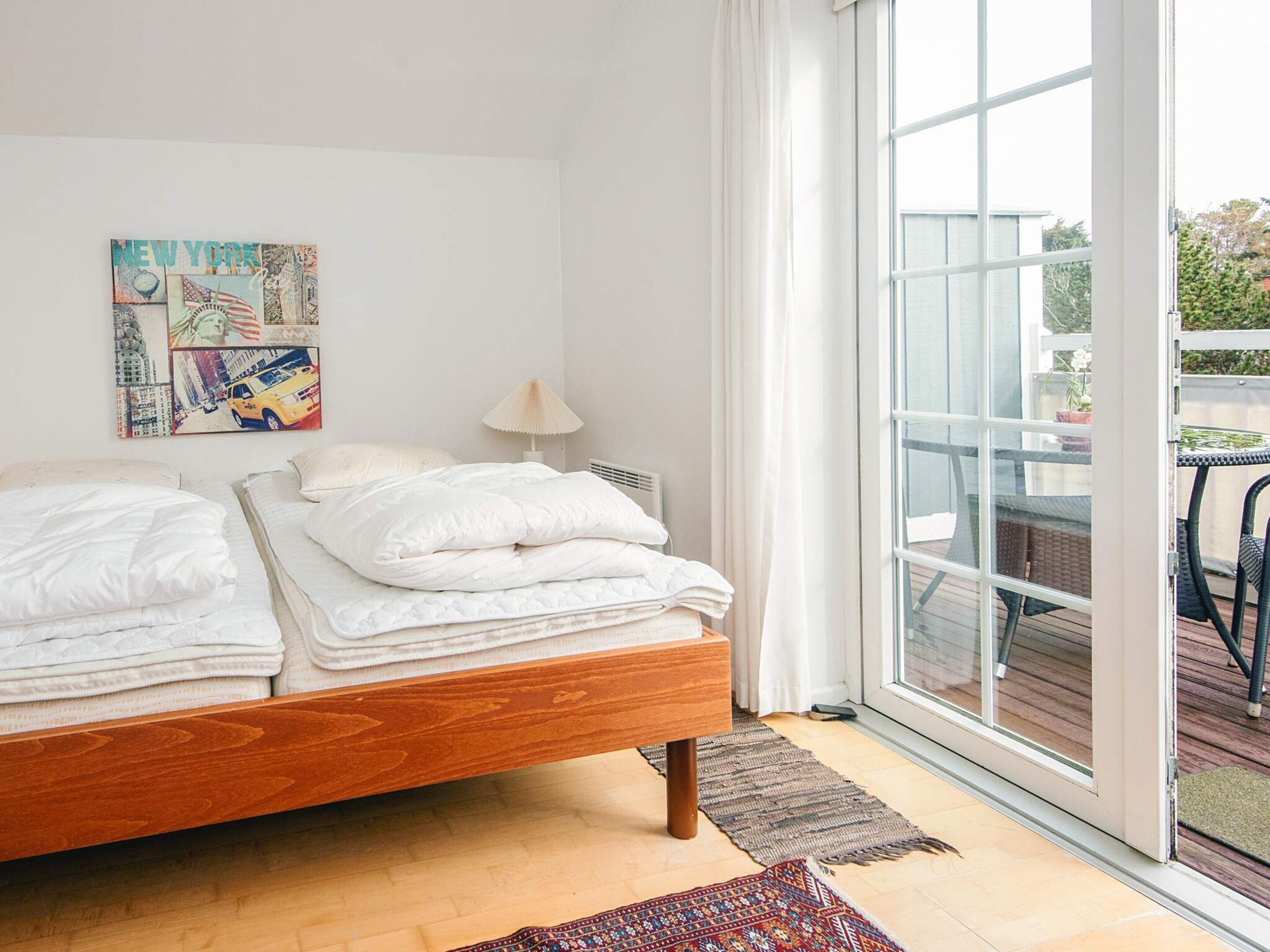 Une chambre avec un lit en bois, du linge de lit blanc, une œuvre d’art représentant un paysage urbain, une lampe et une grande fenêtre donnant sur l’extérieur.