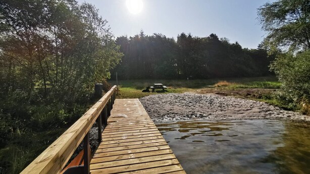 A wooden boardwalk leading to a picnic table by a river, surrounded by trees.
