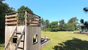 A wooden playhouse with a ladder and windows, surrounded by a grassy yard and trees.