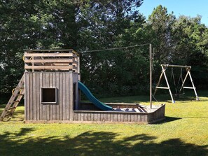 A wooden playhouse with a slide and a swing set in a grassy area.