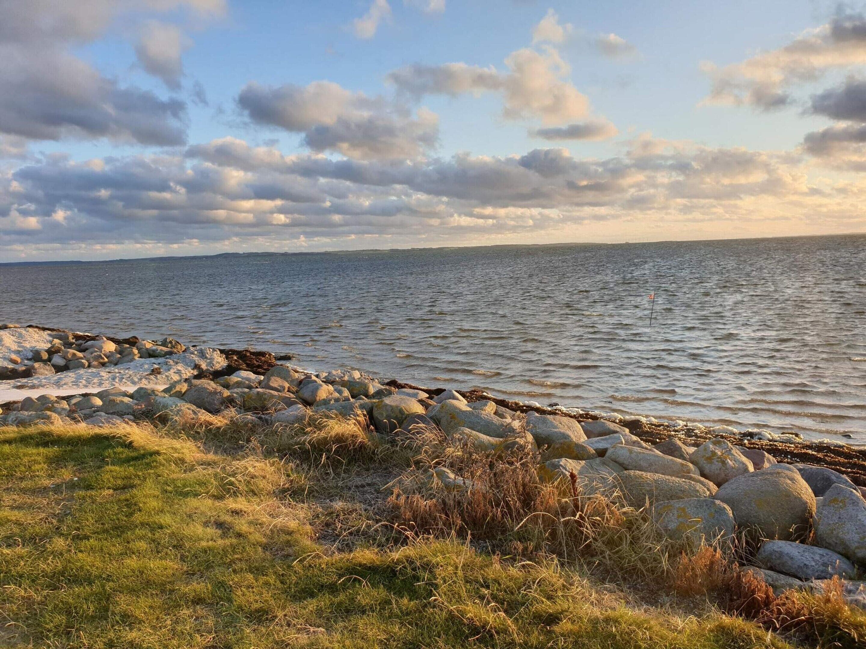 A coastal landscape with a rocky shoreline, grassy areas, and a calm sea under a partly cloudy sky.